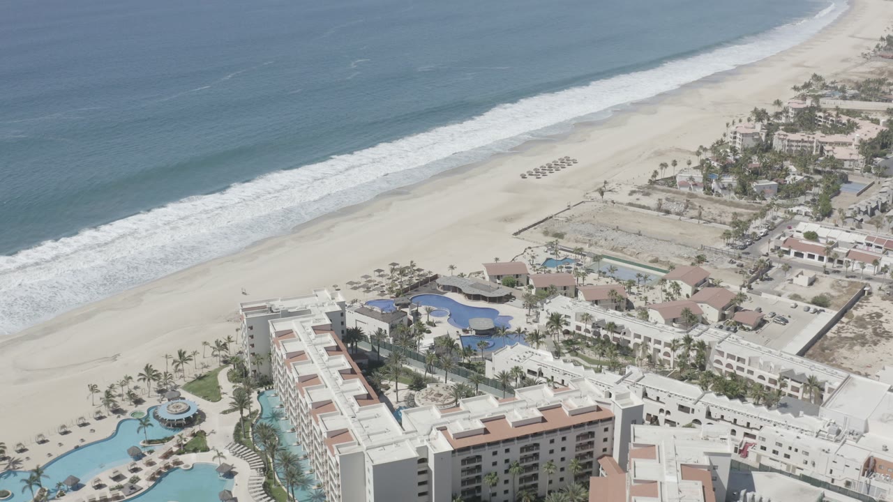 High oblique view in San Jose Del Cabo with hotel roofs lower in frame, pools and palms in the middle, and white surf rolling along the beach, with no visible people.