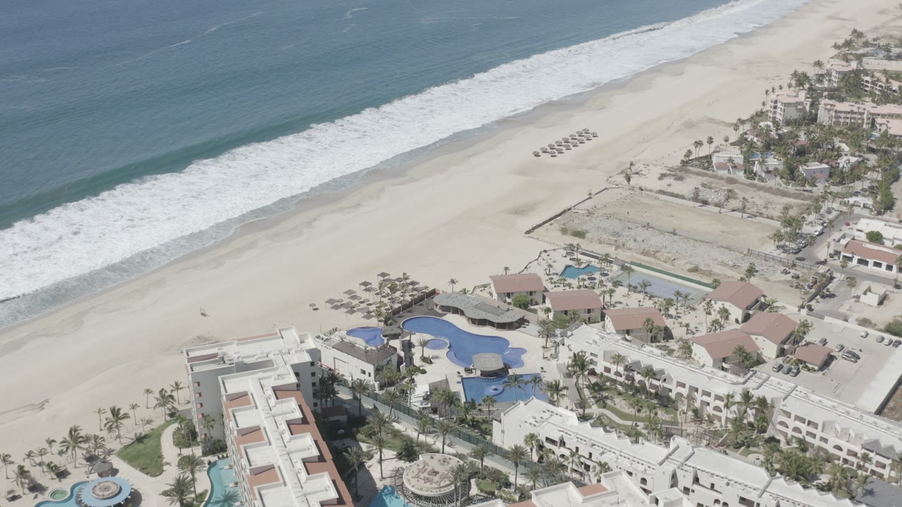 Aerial frame over San Jose del Cabo, Mexico, showing blue ocean, white surf, a wide sandy beach, palm-lined pools, low-rise buildings, and no visible people at the start of the clip.