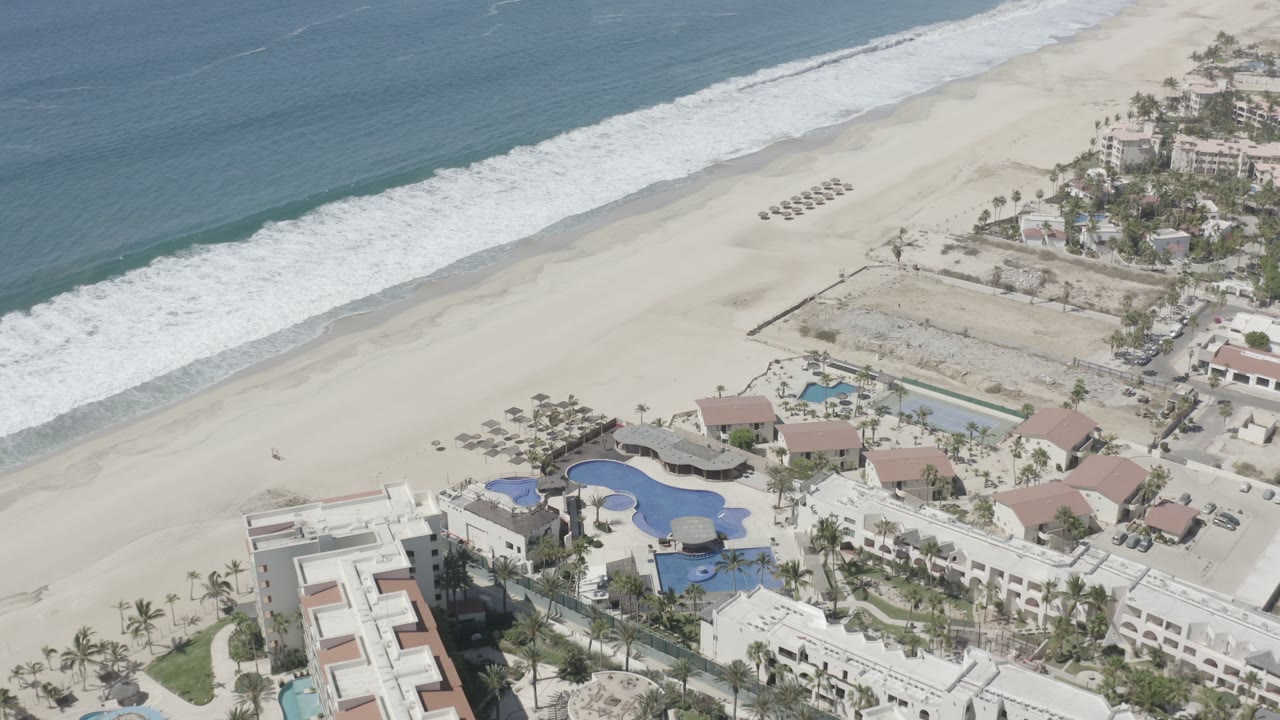 Aerial coastline view with beach umbrellas, surf, buildings, and pools.