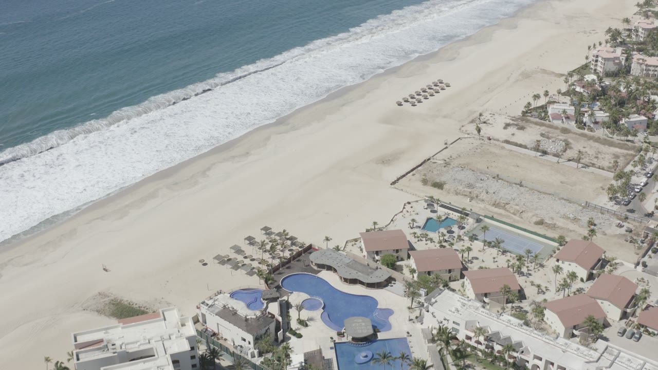 Aerial view of resort pools beside a wide beach and ocean surf.