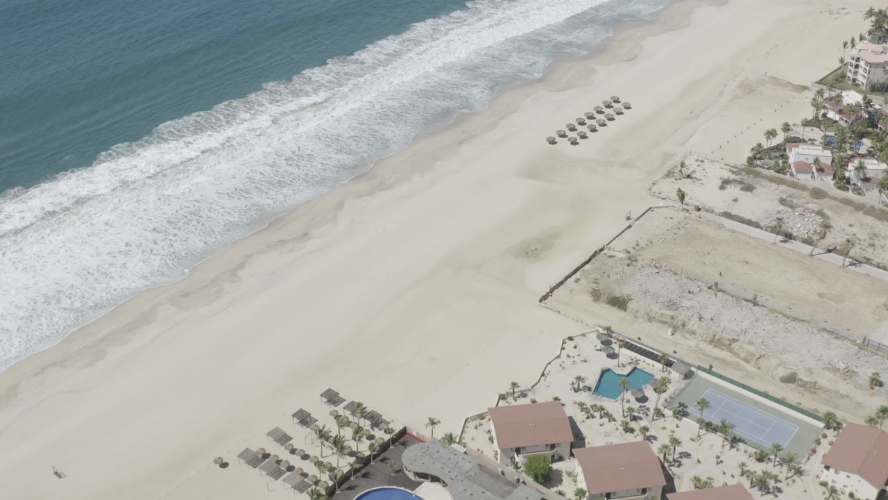 Aerial view over San Jose Del Cabo showing turquoise water, a band of white surf, pale sand, rows of umbrellas, and beach houses at the edge, with no visible people.