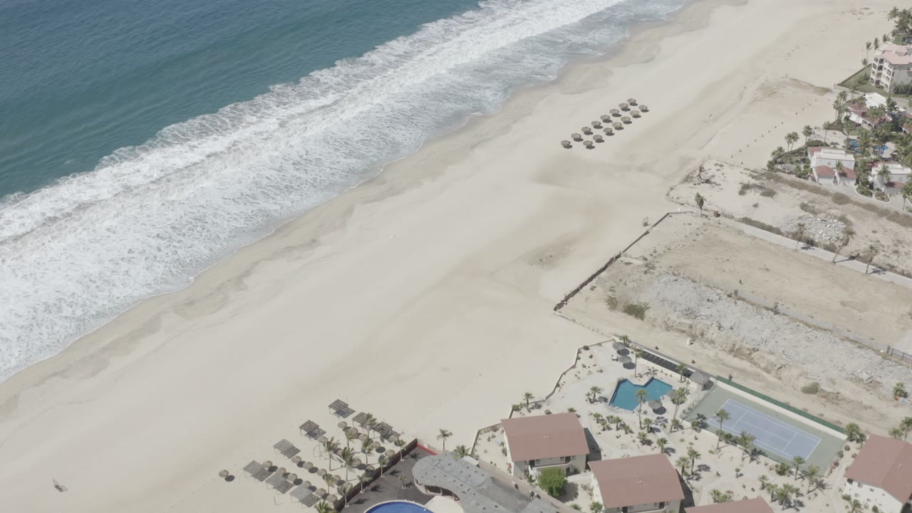 Aerial frame from San Jose Del Cabo with surf sliding diagonally along the beach, umbrella rows near the upper sand, and homes and recreational courts along the lower edge, with no visible people.
