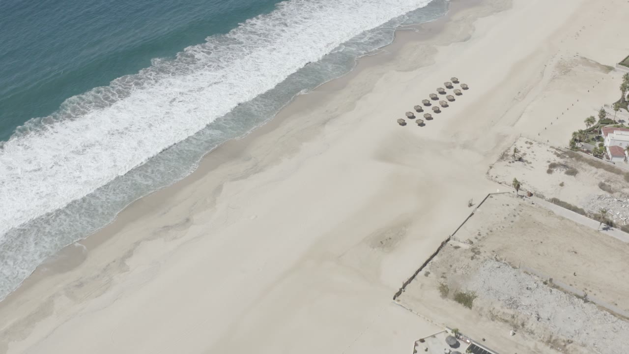 Overhead view of a San Jose Del Cabo beach with blue-green ocean, white surf, pale sand, clustered shade structures, and no visible people.