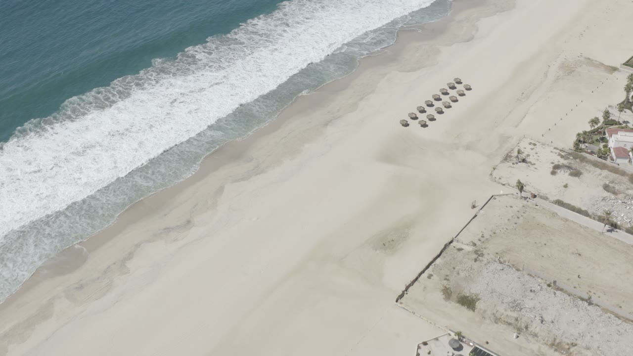 Aerial view over San Jose Del Cabo sand where a white surf line borders the beach and a small cluster of shade structures sits inland, with no visible people.