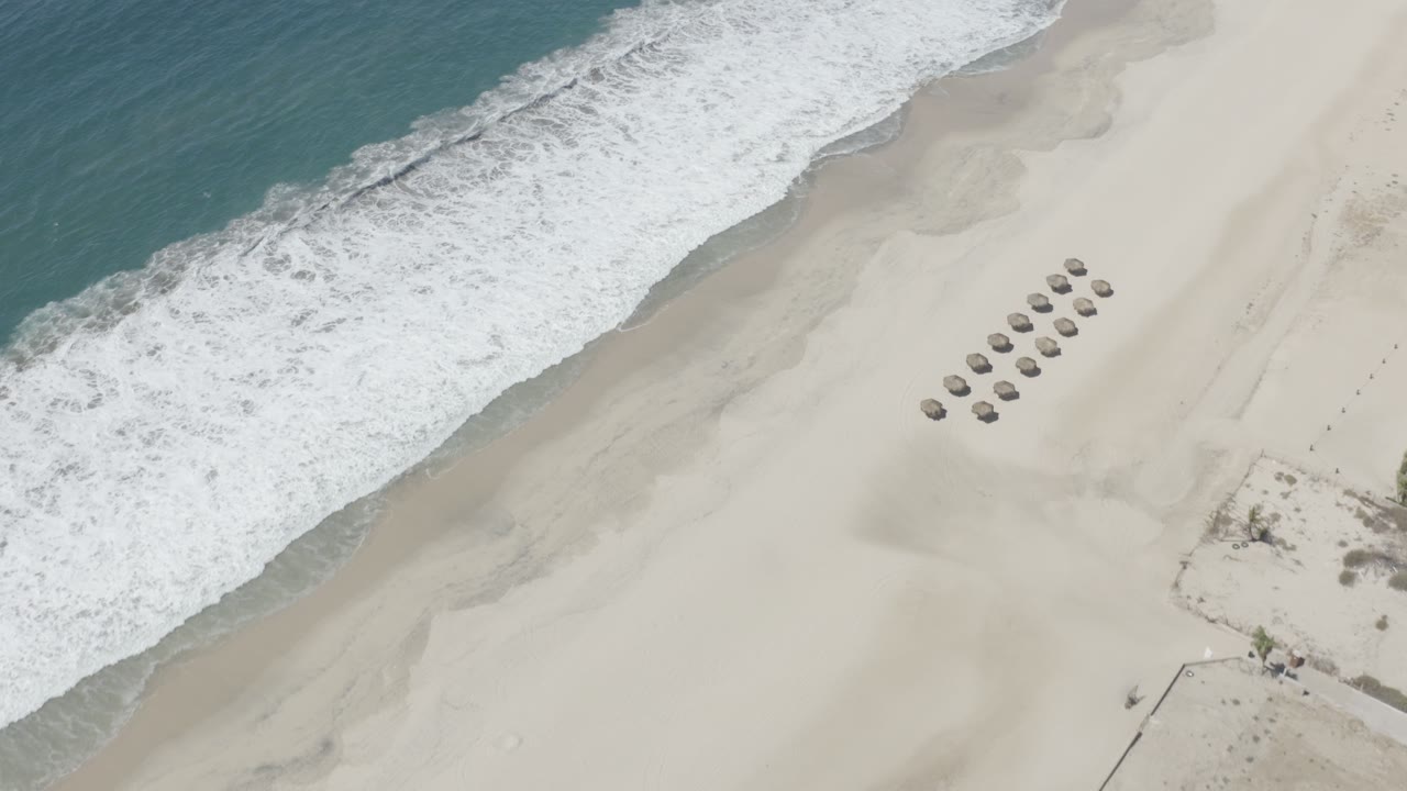 Top-down aerial frame over a San Jose del Cabo beach, with turquoise water and white surf along pale sand, a cluster of round shade structures to the right, and no visible people.