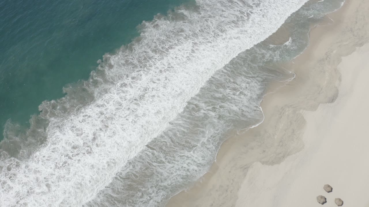 Overhead view in San Jose Del Cabo, Mexico, with teal ocean at left, white seafoam crossing the center, pale beach at right, small rocks near the lower edge, and no visible people.