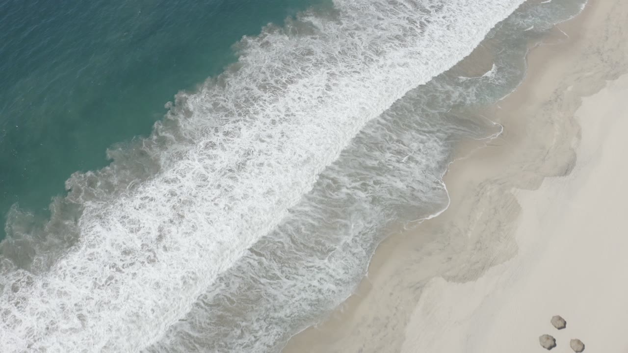 Aerial shoreline frame from San Jose Del Cabo, Mexico, showing a broad band of white foam over shallow water, pale sand to the right, teal water to the left, and no visible people.