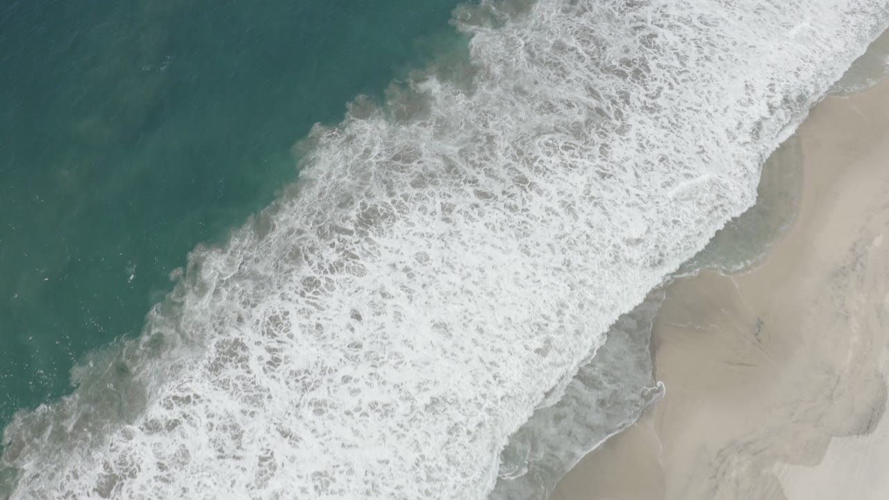 Top-down aerial frame of white surf crossing from the upper right toward pale sand in San Jose del Cabo, Mexico, with teal water at left and no visible people.