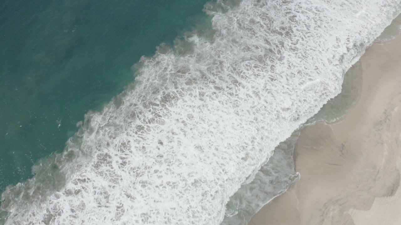 Aerial view of dense white wave foam sliding across the sandy shoreline in San Jose del Cabo, Mexico, with deeper teal water on the left and no visible people.