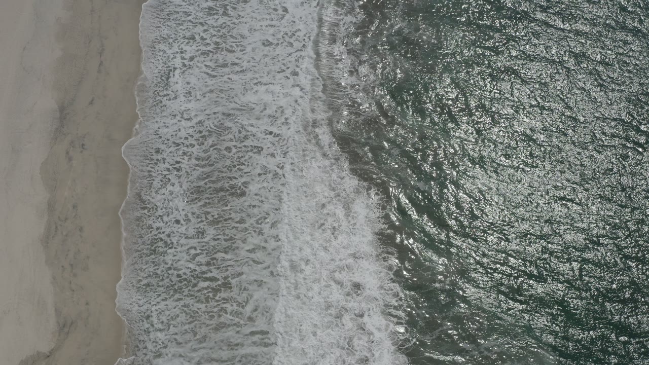 Top-down view of San Jose del Cabo surf with white foam crossing pale sand and dark green water sparkling on the right, with no visible people.