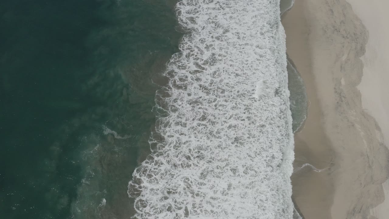 Overhead view of dark green ocean and white surf meeting pale sand in San Jose Del Cabo, Mexico, with no visible people.