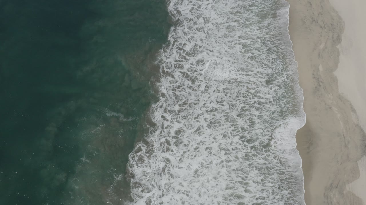 Top-down view of San Jose del Cabo surf with dark teal water on the left, white foam across the center, pale sand on the right, and no visible people.