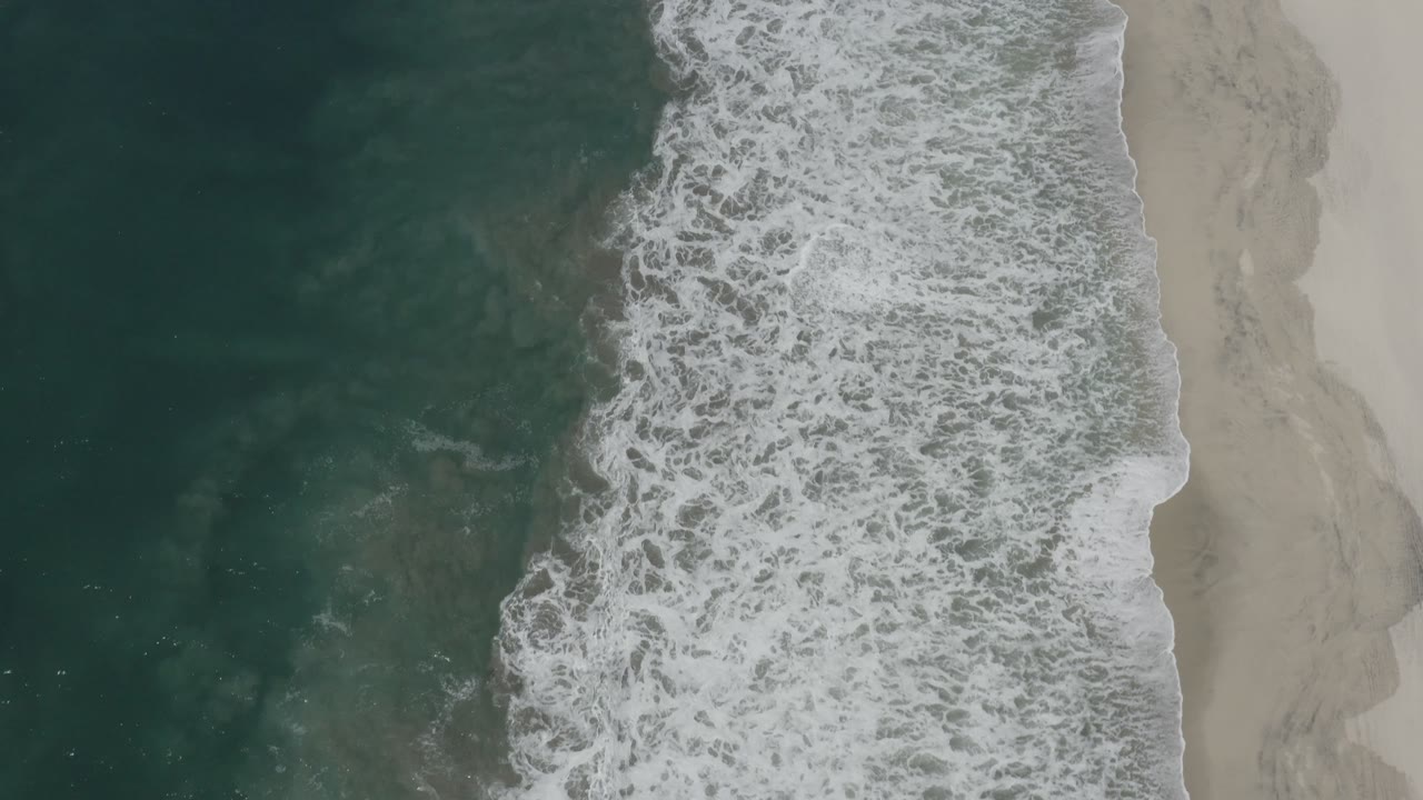 Aerial frame over San Jose del Cabo showing the surf band shifted slightly along the shore, with textured teal water, white breakers, pale sand, and no visible people.