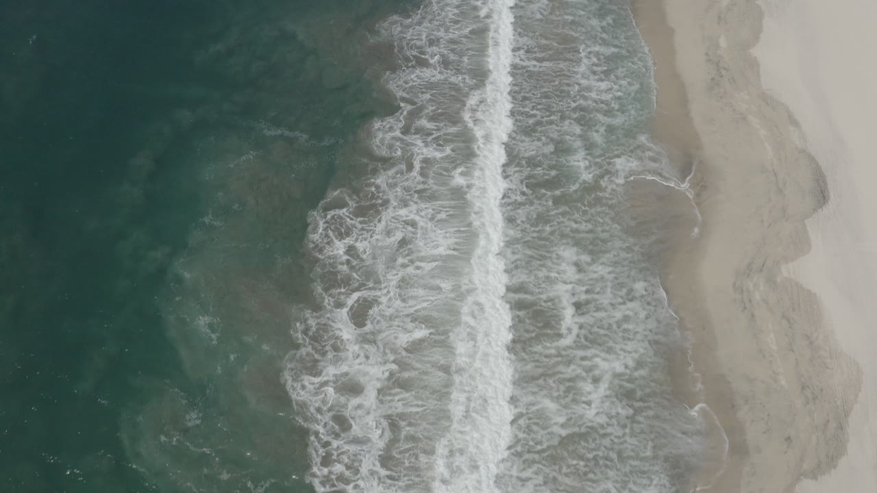 Bird's-eye view of teal ocean, white breaking surf, and pale sand in San Jose del Cabo, Mexico, with no visible people at the opening moment.