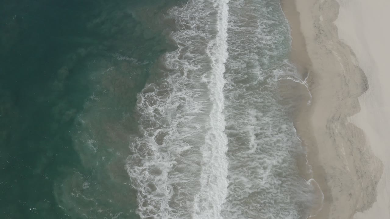 Aerial frame looking straight down at a narrow white surf band between dark teal water and pale beach in San Jose del Cabo, Mexico, with no visible people.