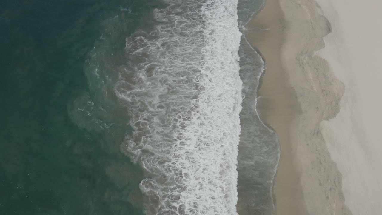 Overhead view of San Jose Del Cabo surf breaking between dark green water and pale sand, with no visible people.