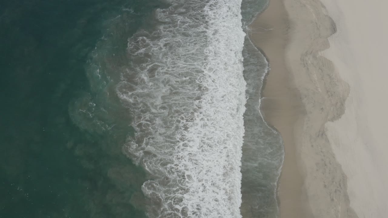 A foamy wave line runs down the center of the San Jose Del Cabo beach frame, with darker water at left, smooth sand at right, and no visible people.