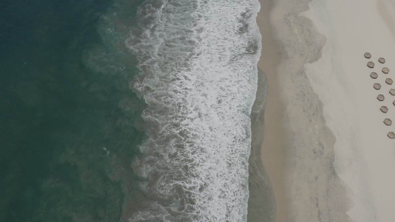 Aerial view of San Jose del Cabo surf breaking along pale beach, with dark teal water on the left, a narrow wet sand band, round umbrellas at right, and no visible people.