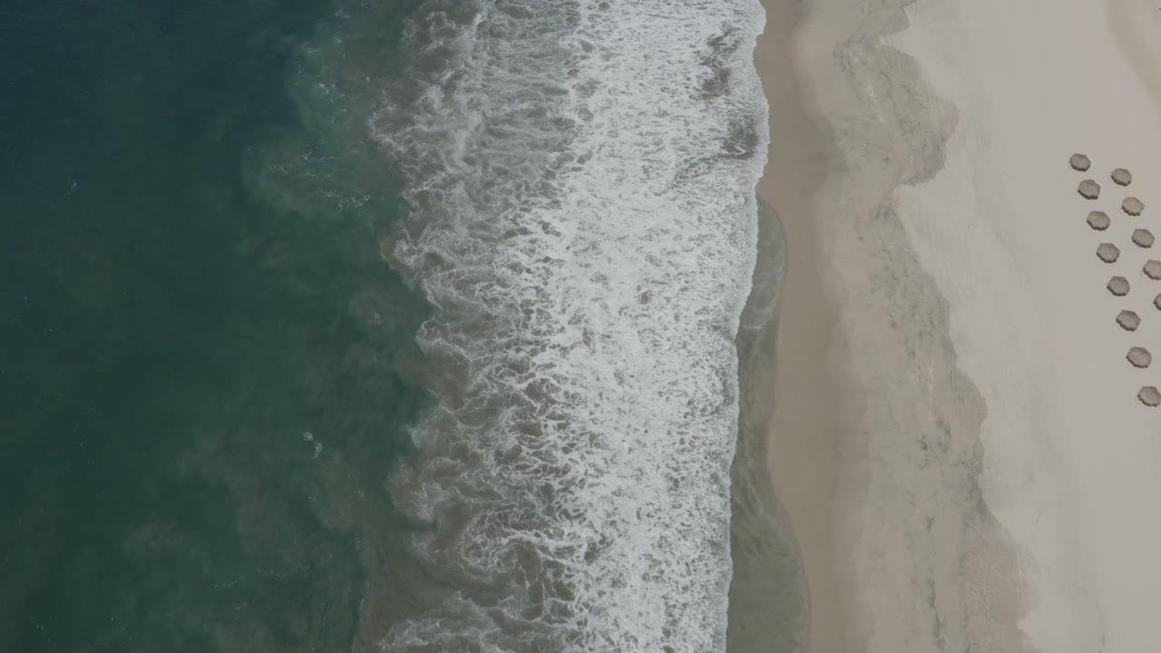Top-down San Jose del Cabo beach frame with foamy water pushing farther across the center shoreline, dry sand to the right, small round umbrellas near the upper right, and no visible people.