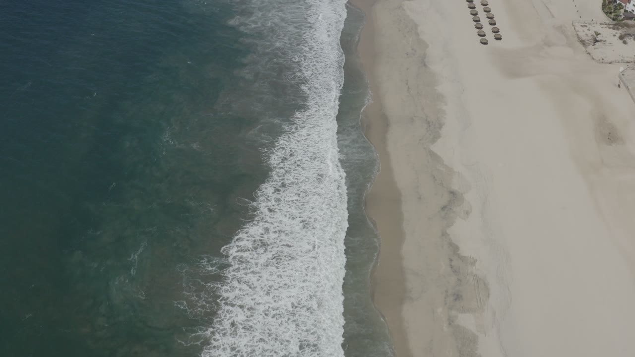 High overhead view of San Jose del Cabo shoreline with dark teal water, a vertical band of white surf, pale sand, and rows of beach loungers near the top edge.