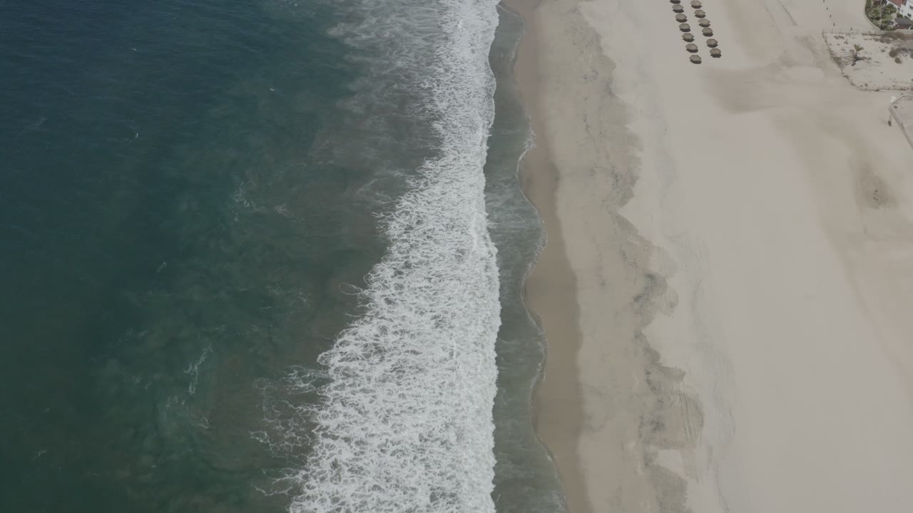 Aerial shoreline with foamy waves beside broad empty sand.