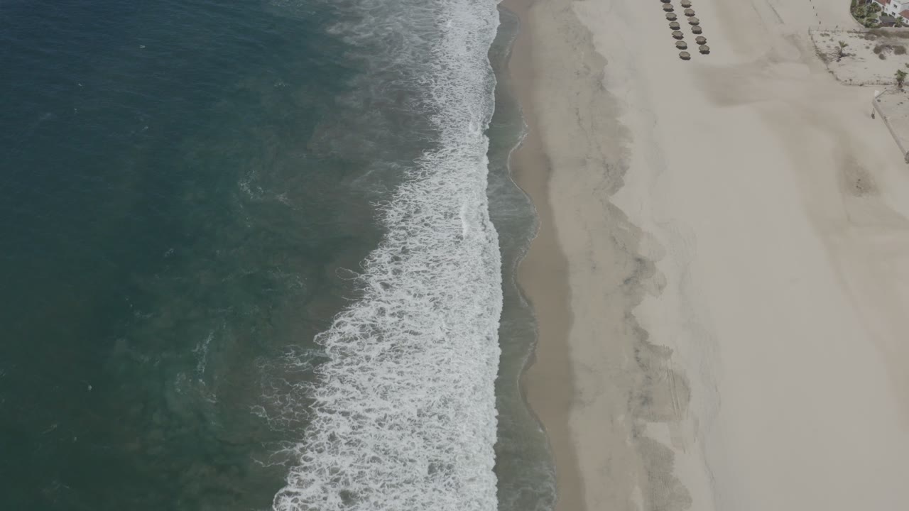 Top-down view of the San Jose del Cabo coast with deeper teal water, a thick white surf band, pale sand, and the beach setup cropped near the top right.