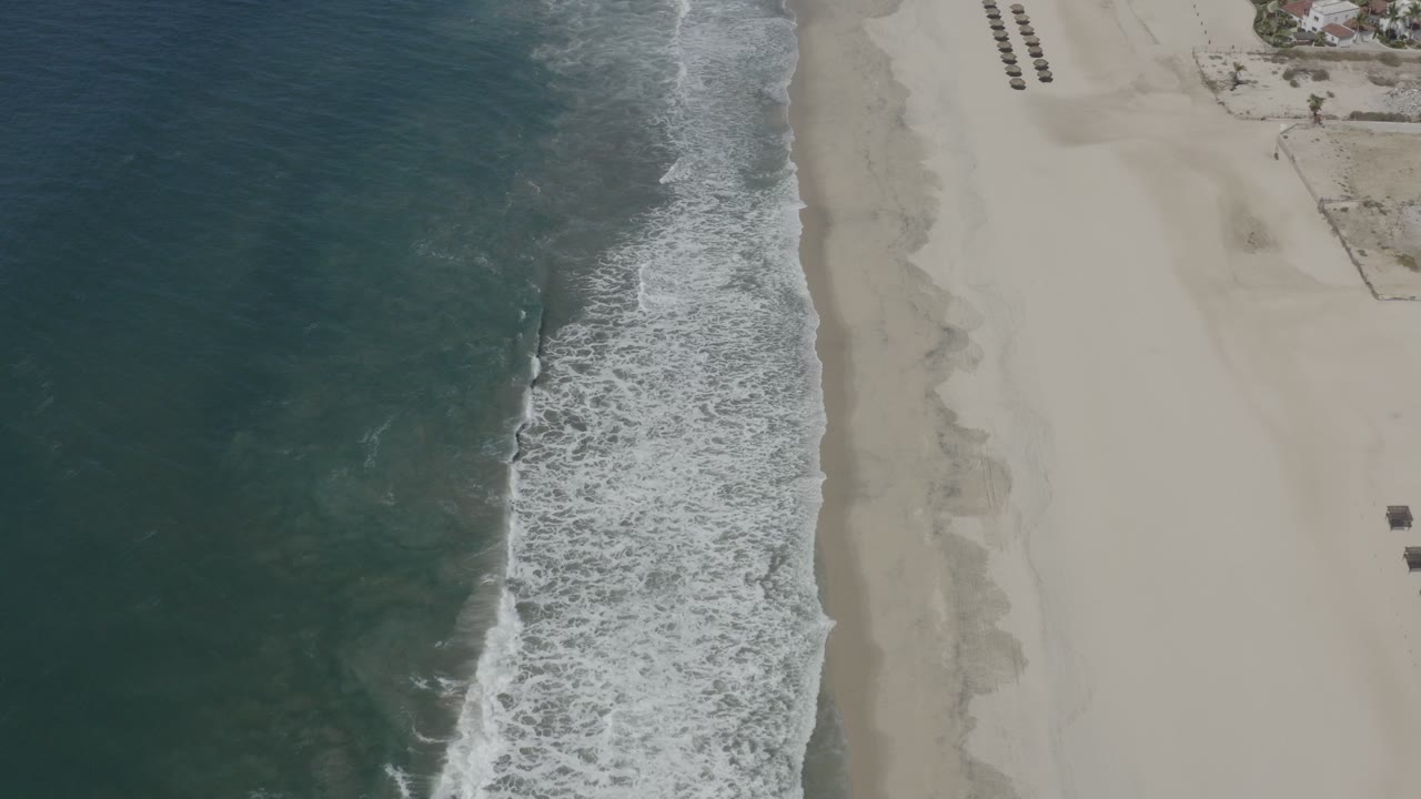 Overhead view of San Jose del Cabo shoreline with teal ocean at left, white surf along pale sand, and no visible people at the start of the clip.