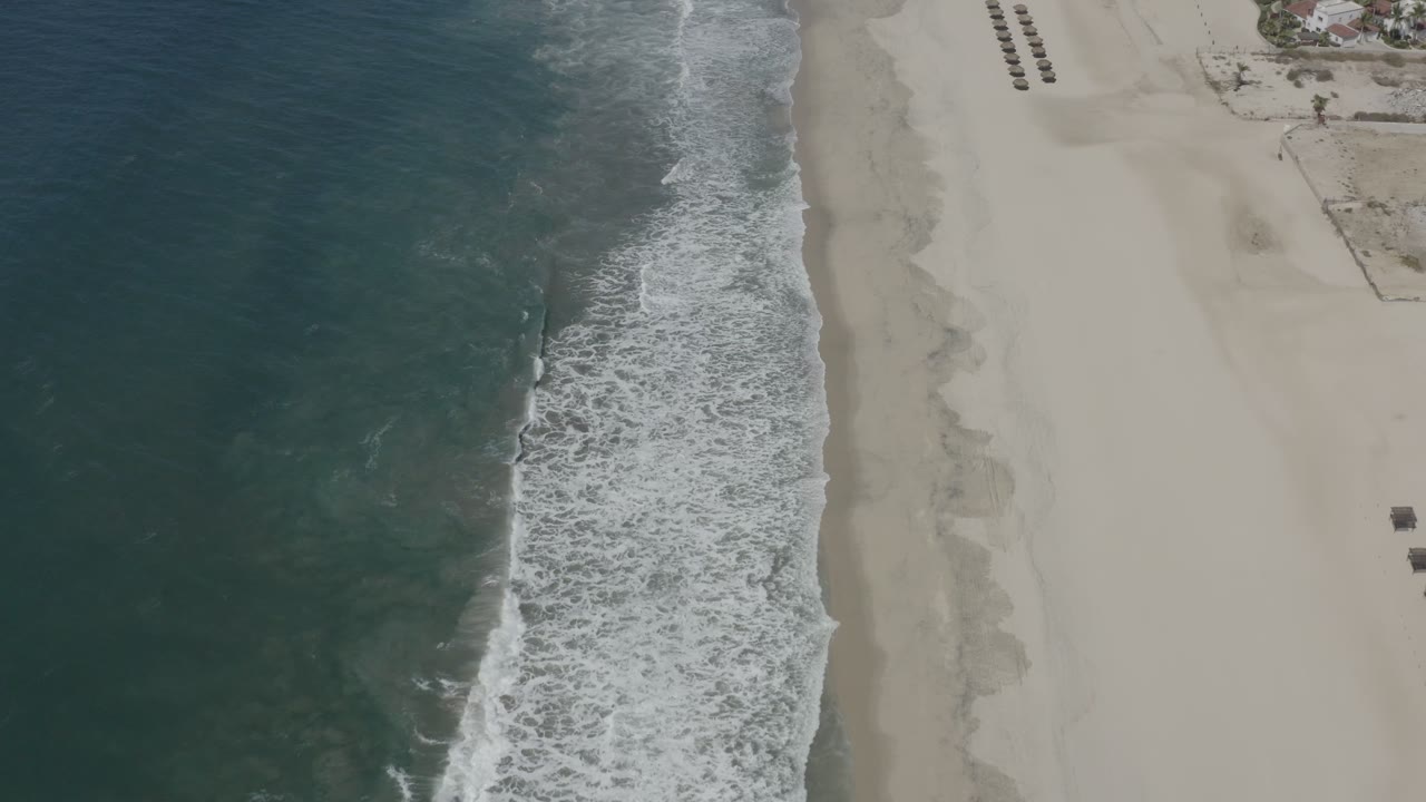 Aerial view of San Jose del Cabo beach where a wide band of foam reaches the sand and the beach remains mostly empty with no visible people.