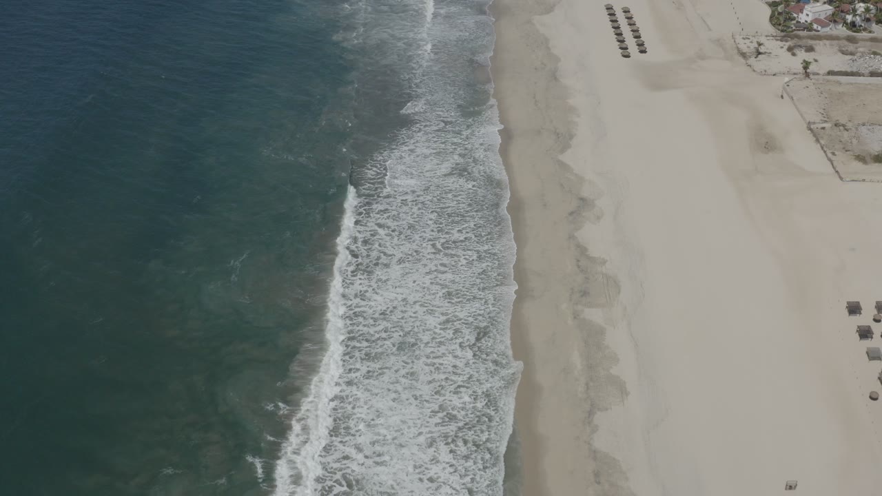 Drone view over San Jose del Cabo coastline with darker water on the left, a foamy surf band near center, pale sand on the right, and no visible people.