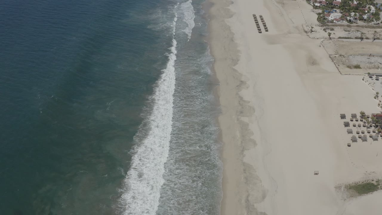 Aerial shoreline frame in San Jose del Cabo showing the foamy wave band closer to center, broad empty sand to the right, and small beach furniture clusters beyond it.