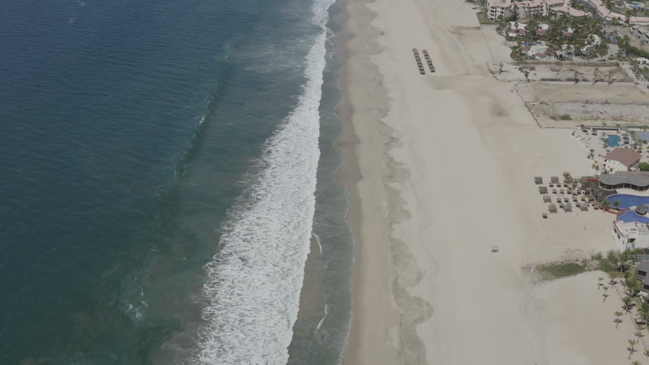 High aerial view of San Jose Del Cabo shoreline with blue ocean on the left, white surf on pale sand, resort structures at the upper right, and no visible people.