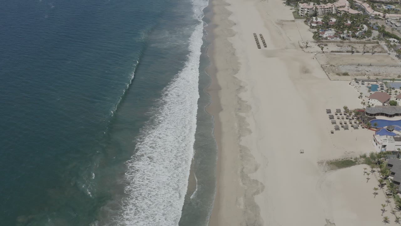 Drone-style view of surf rolling toward a pale beach with buildings at the edge.