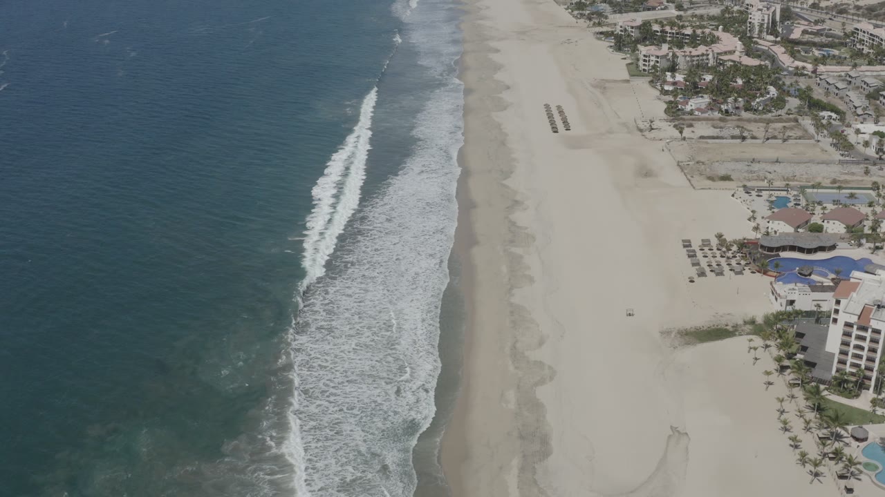 High aerial frame in San Jose del Cabo, Mexico, showing a long band of breaking surf, open sand, and residential buildings with pools along the beach edge.