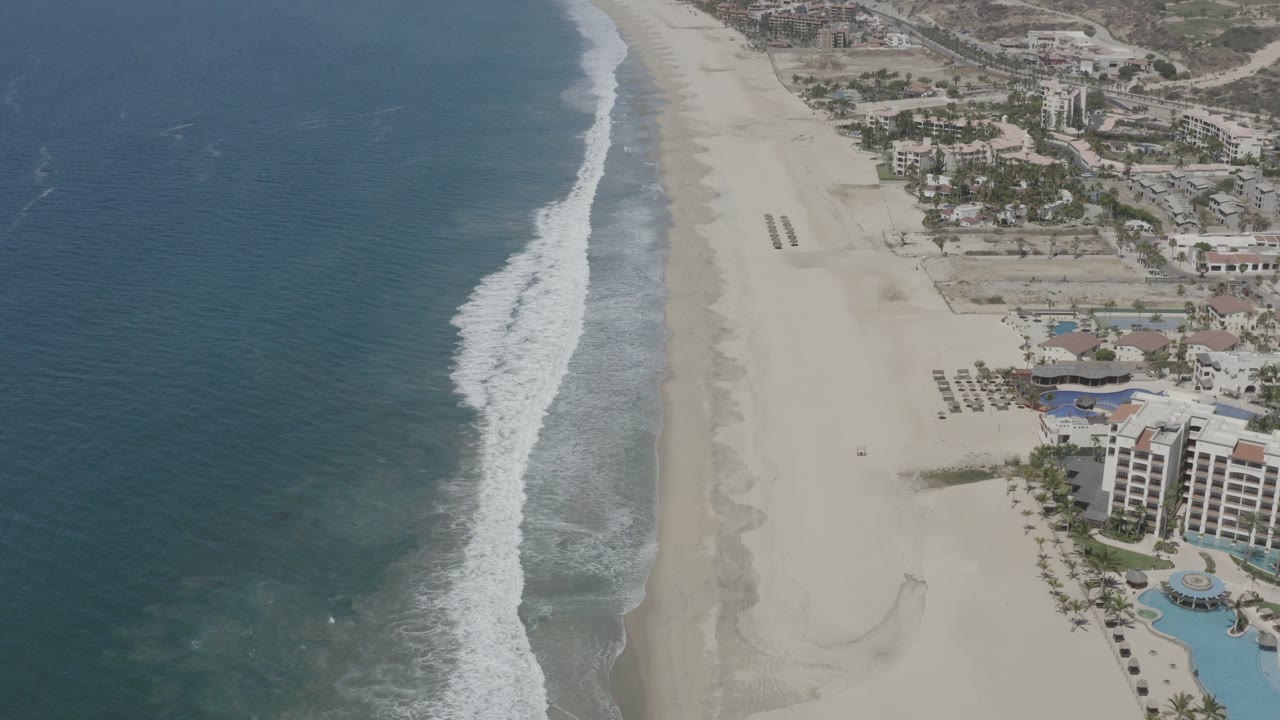 Aerial coastline with ocean, surf, sand, resort buildings, and inland development.