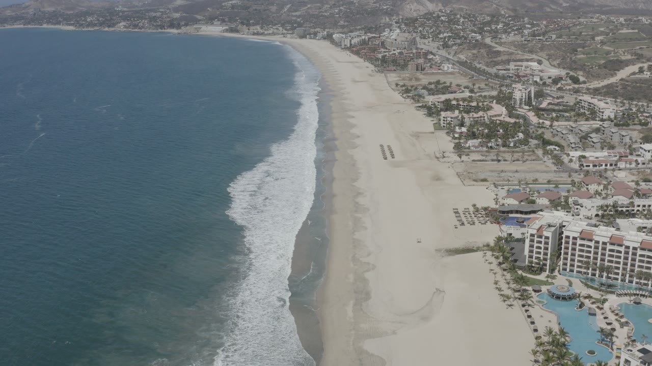 Aerial coastline with blue ocean, sandy beach, surf, and beachfront buildings.