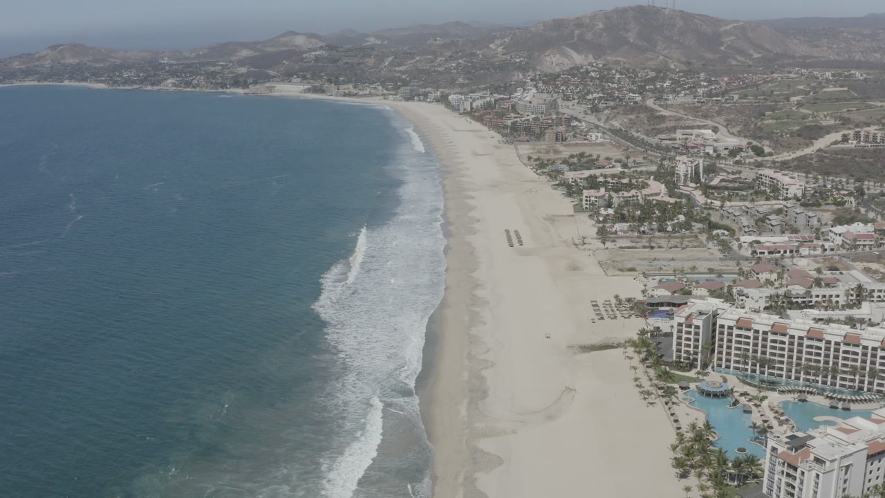 Opening aerial frame over San Jose Del Cabo showing blue ocean, a broad sandy beach, beachfront resort buildings, hazy mountains, and no visible people.
