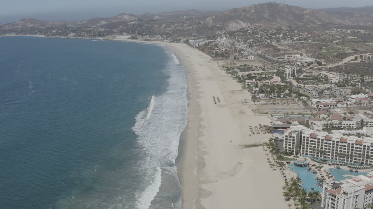Aerial view along the San Jose Del Cabo beach with a longer band of white surf, pale sand, hotel blocks, turquoise pools, distant hills, and no visible people.