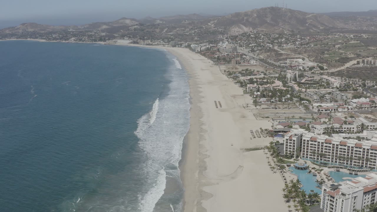 Coastal aerial with ocean to the left, sandy shoreline, resort buildings, and hills.