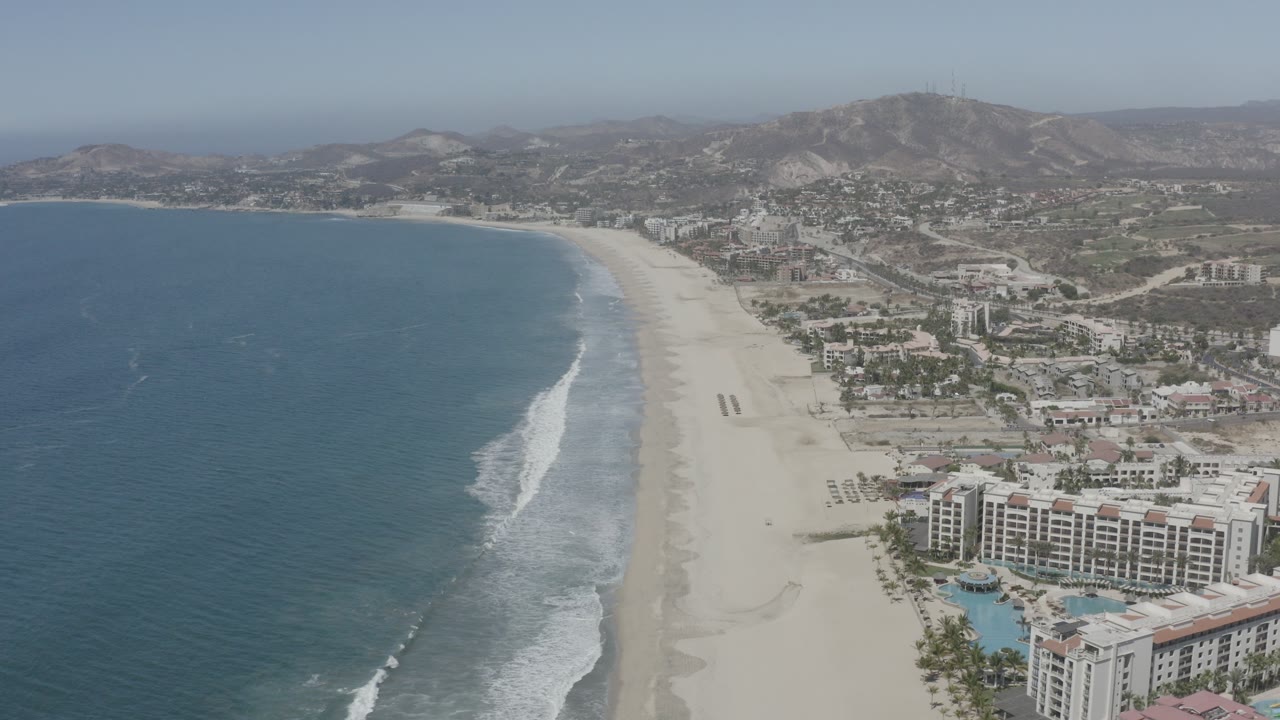 Aerial view over San Jose del Cabo with blue ocean at left, pale beach and surf along the center, resort buildings at lower right, inland roads, dry hills, and no visible people.