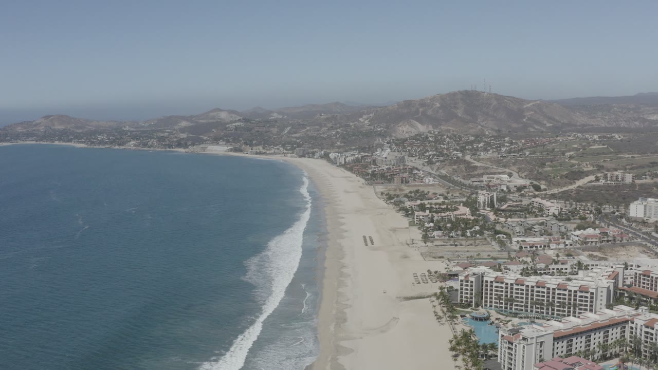Aerial view over San Jose del Cabo, Mexico, with blue ocean at left, a wide sandy beach, beachfront buildings in the lower right, distant town and mountains, and no visible people.