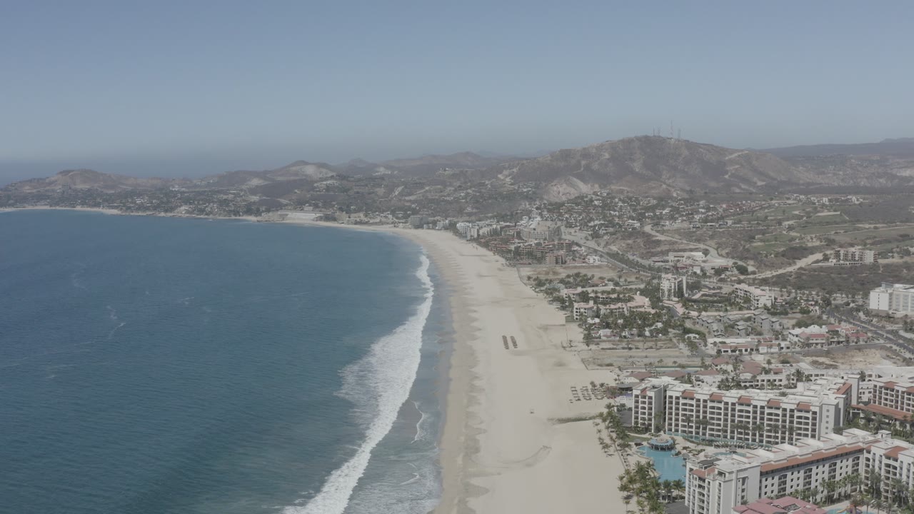 Aerial beach view with ocean, surf, resort-like buildings, town, and dry hills.