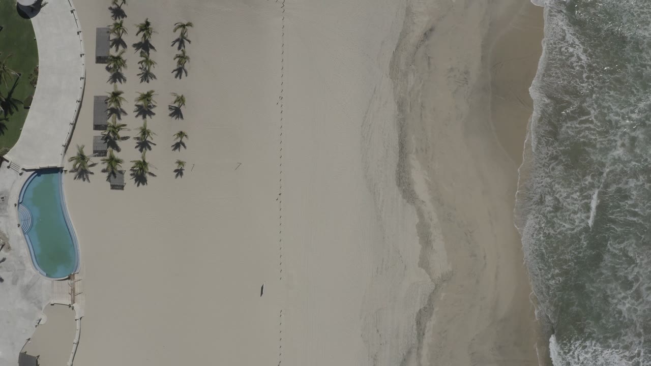 Aerial view over San Jose Del Cabo shoreline with surf washing along the right edge, open sand in the center, and poolside landscaping at left, with no visible people.