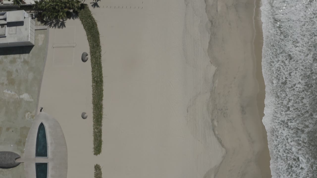 Overhead San Jose del Cabo shoreline frame showing the surf band shifted along the sand beside landscaping and an oval pool, with no visible people.