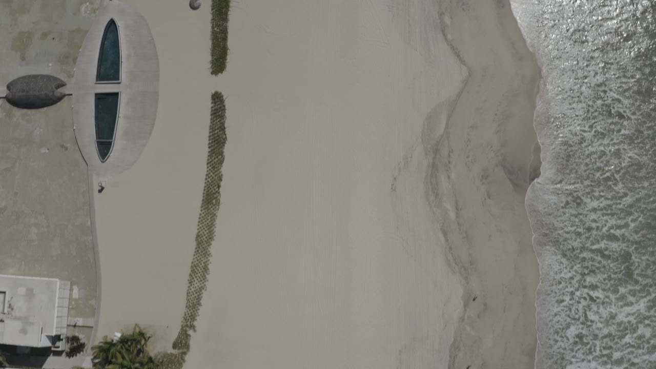 Top-down beachfront with surf, sand, oval pool or window feature, and a small building edge.