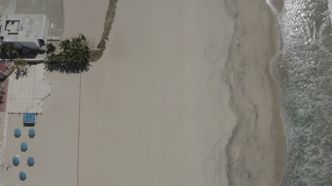 Aerial frame in San Jose del Cabo with blue beach umbrellas near a resort boundary, a broad stretch of pale sand, and bright wave wash along the shoreline, with no visible people.