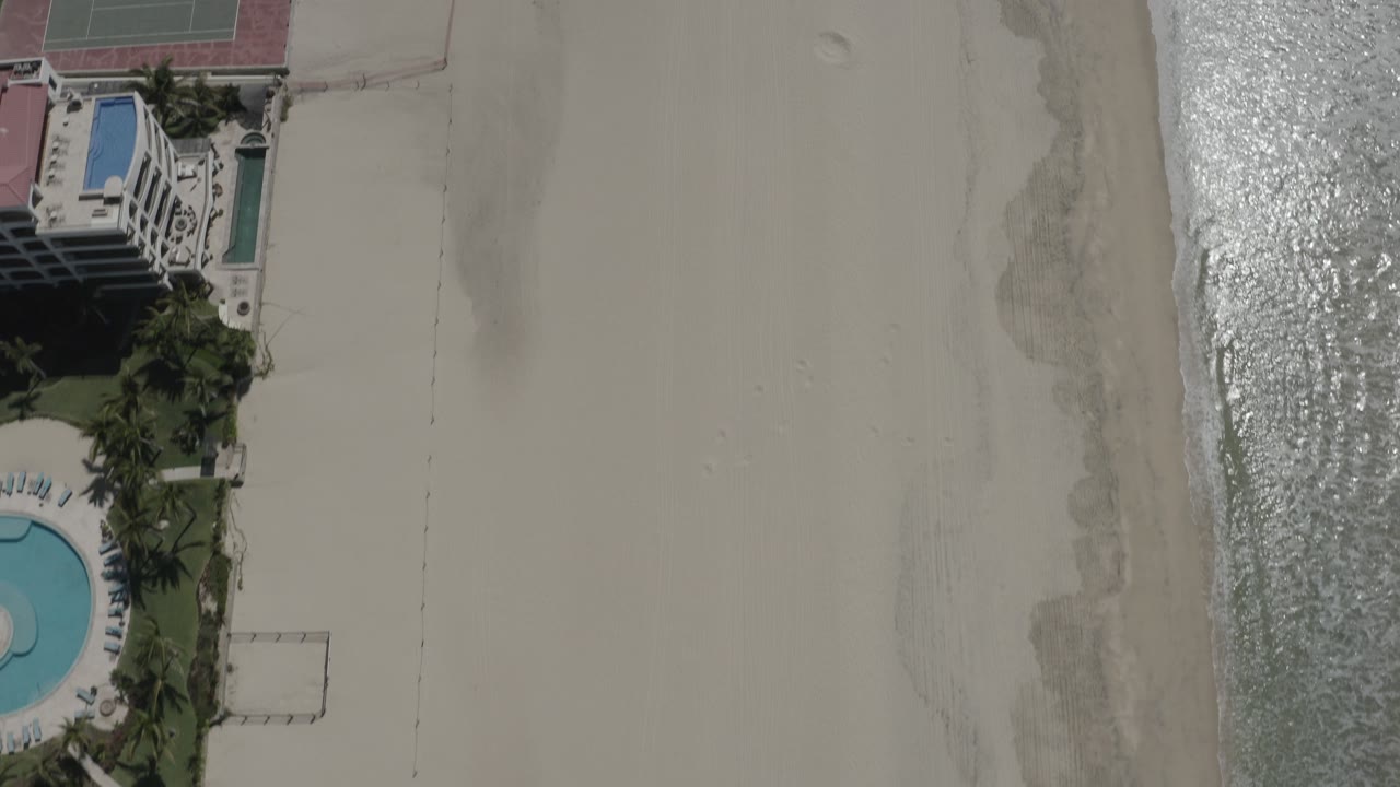 Top-down aerial view of beach, surf, palms, pools, and a waterfront building.