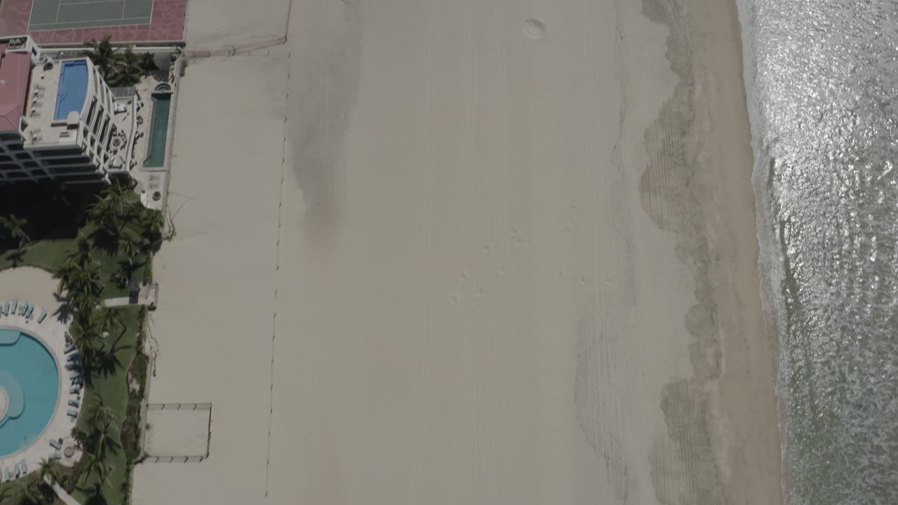 Aerial overhead frame in San Jose del Cabo, Mexico with the white building and pools higher in frame, empty sand across the center, bright ocean glare at the side, and no visible people.