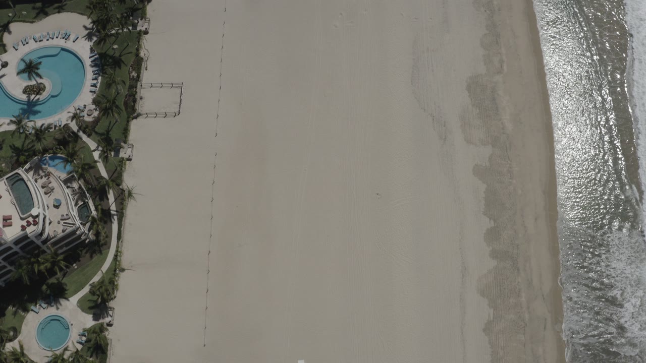 Overhead frame from San Jose del Cabo, Mexico showing beachfront houses with blue pools beside pale sand and a narrow strip of surf, with no visible people.