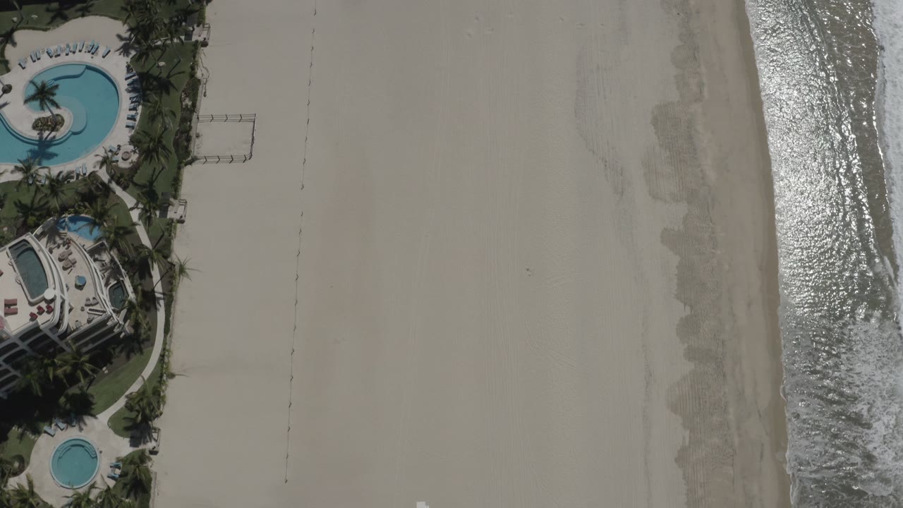 Aerial view in San Jose del Cabo, Mexico with the beachfront property edge at left, pale open sand through the center, and reflective surf along the right side, with no visible people.