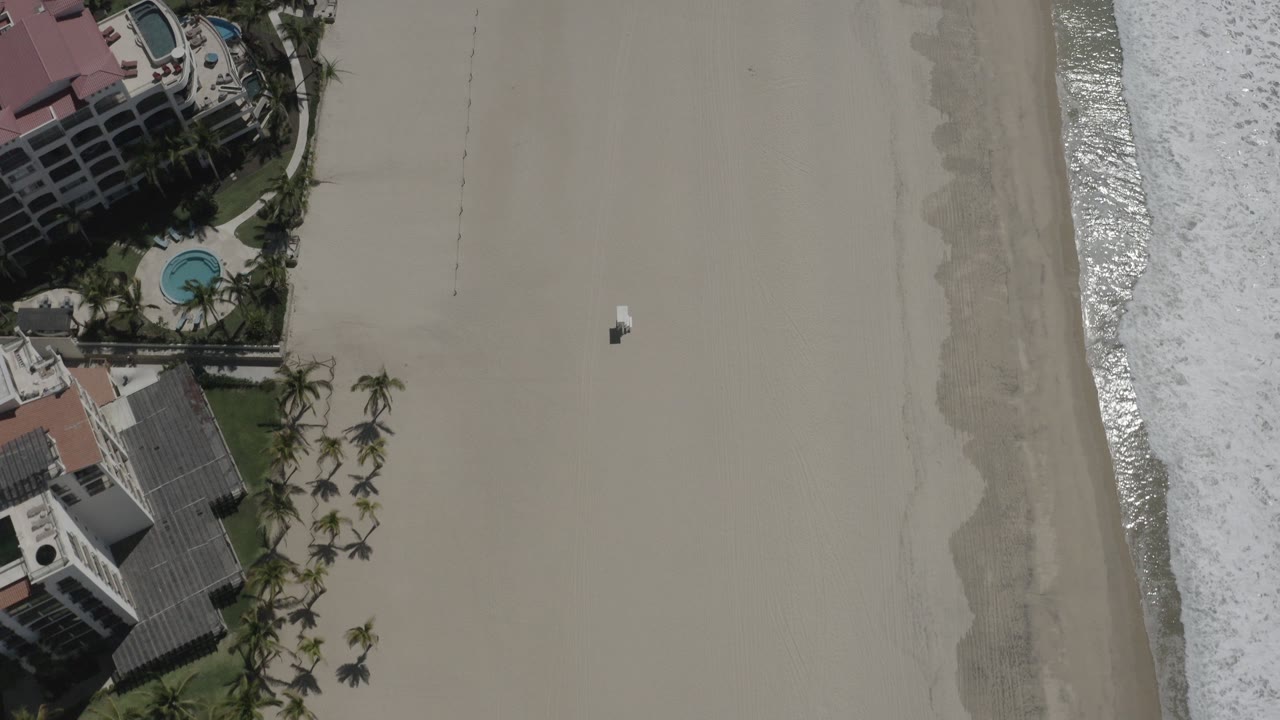 High overhead view of San Jose del Cabo beach with hotel buildings clipped at the left edge, palm landscaping below, surf at the right edge, and no visible people.